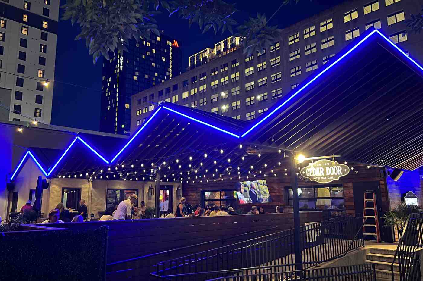 Outdoor restaurant patio at night illuminated with vibrant blue X Series Roofline Lighting, enhancing the modern design, complemented by string lights and a lively dining atmosphere.