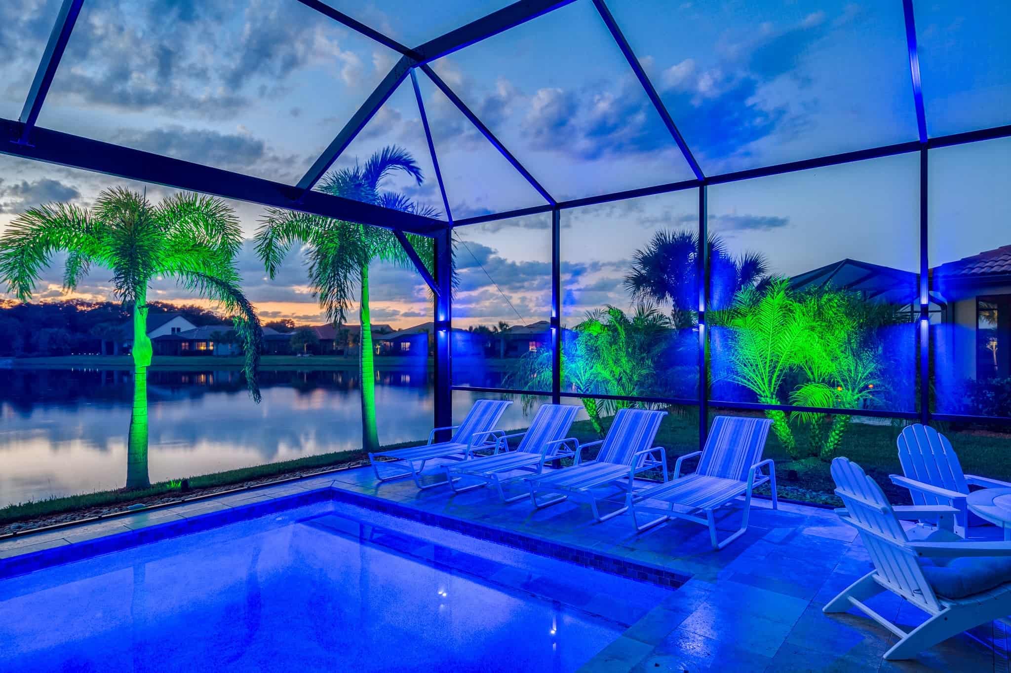 Screened-in pool area with blue and green lighting, overlooking a lake at sunset.