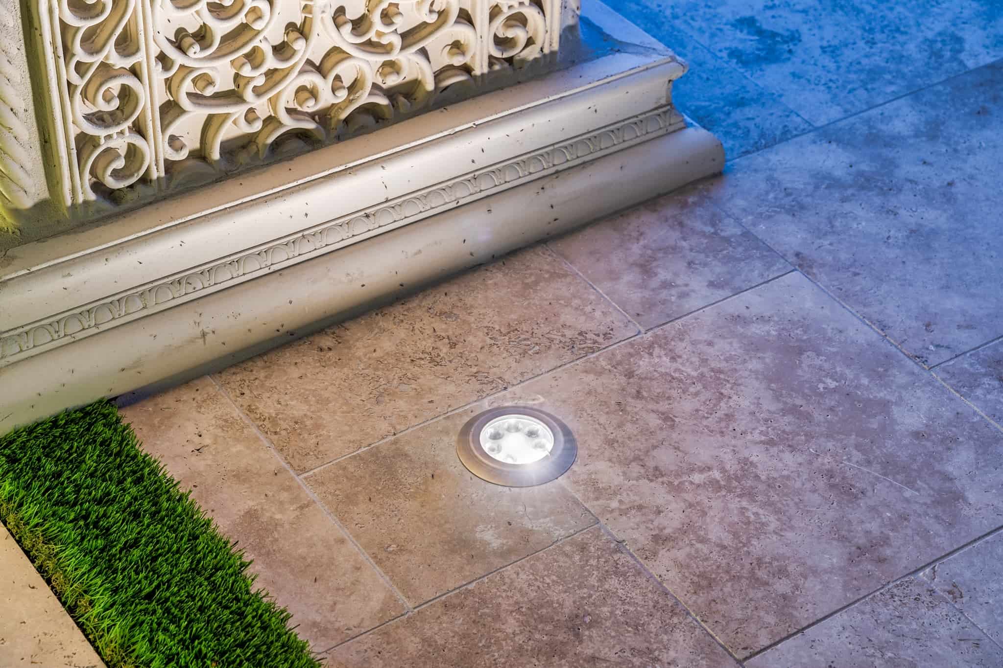 In-ground LED uplight illuminating decorative stone trim on a tiled patio.