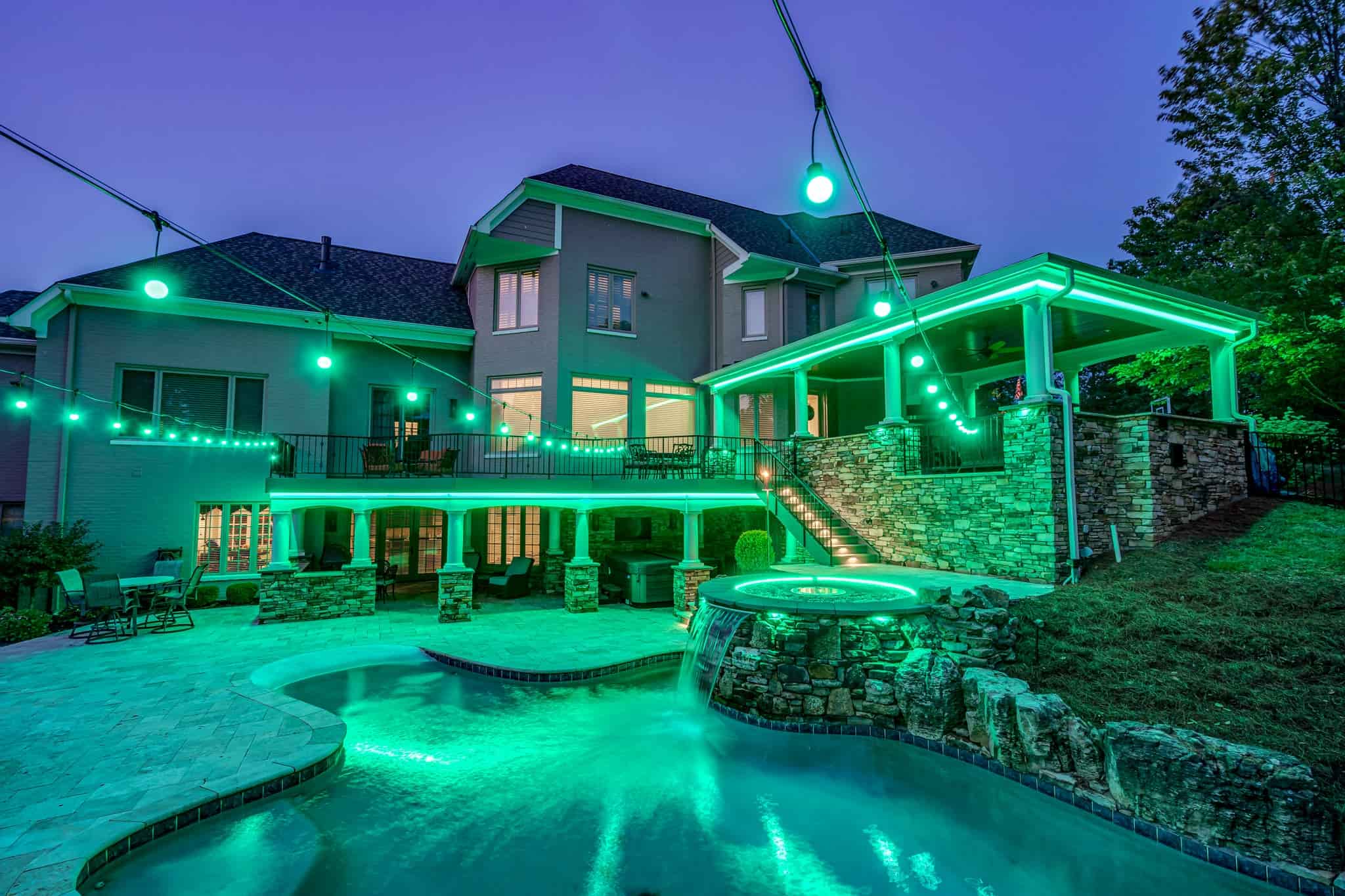 Luxurious backyard with a pool, waterfall, and outdoor living space illuminated by vibrant green landscape and string lighting.