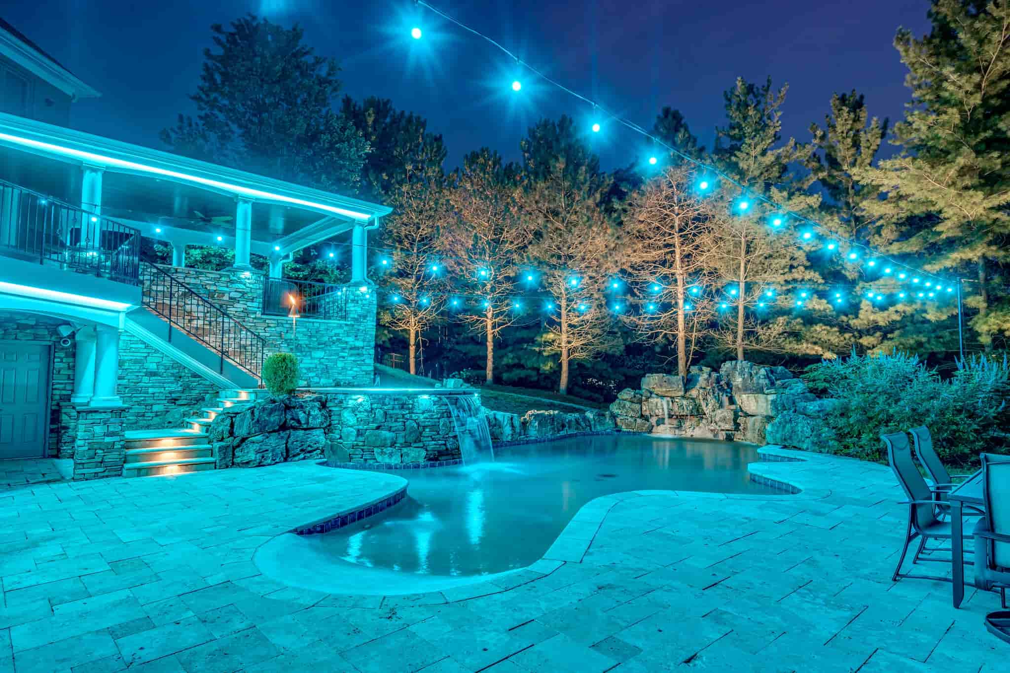 Luxury backyard with a pool, waterfall, and vibrant blue string lighting at night.