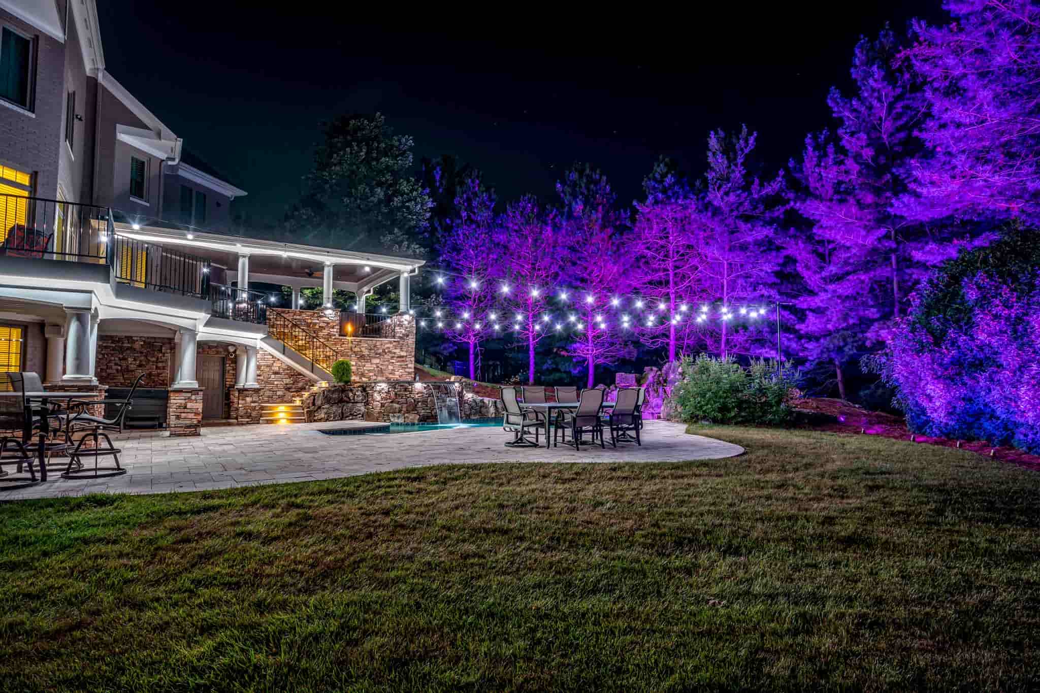 Luxury backyard patio with ambient string lights and vibrant purple landscape lighting at night.